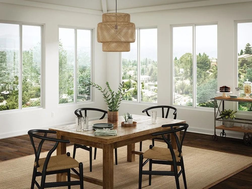 A sunlit dining room with a wooden table and black chairs, surrounded by large windows overlooking a lush landscape.