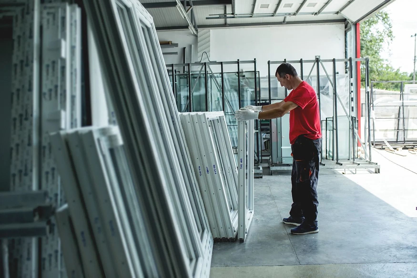 A worker in a warehouse wearing a red shirt inspects window frames. White frames are stacked, ready for use.