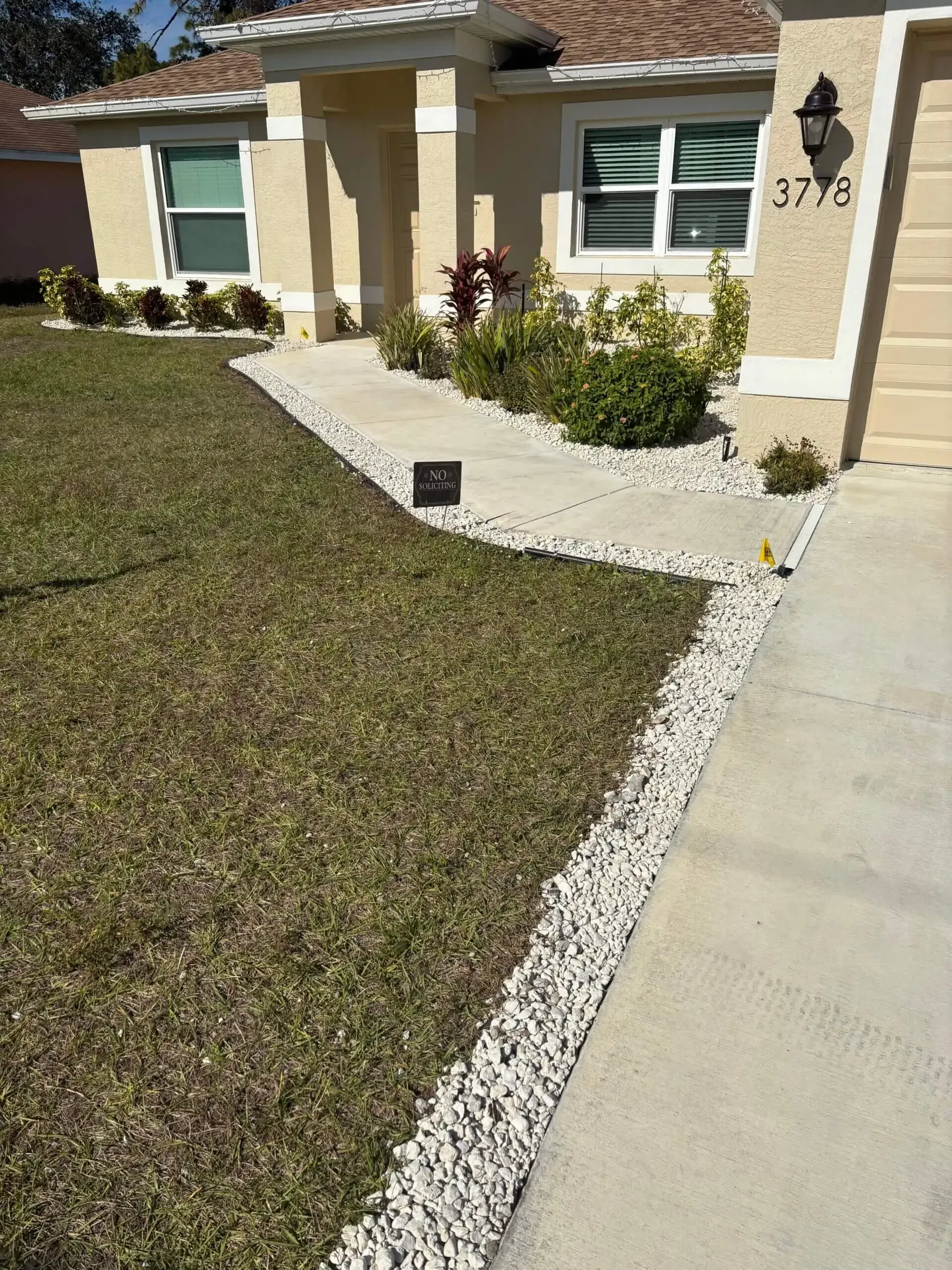 A beige house with a walkway lined with white stones, beside a driveway and lawn.