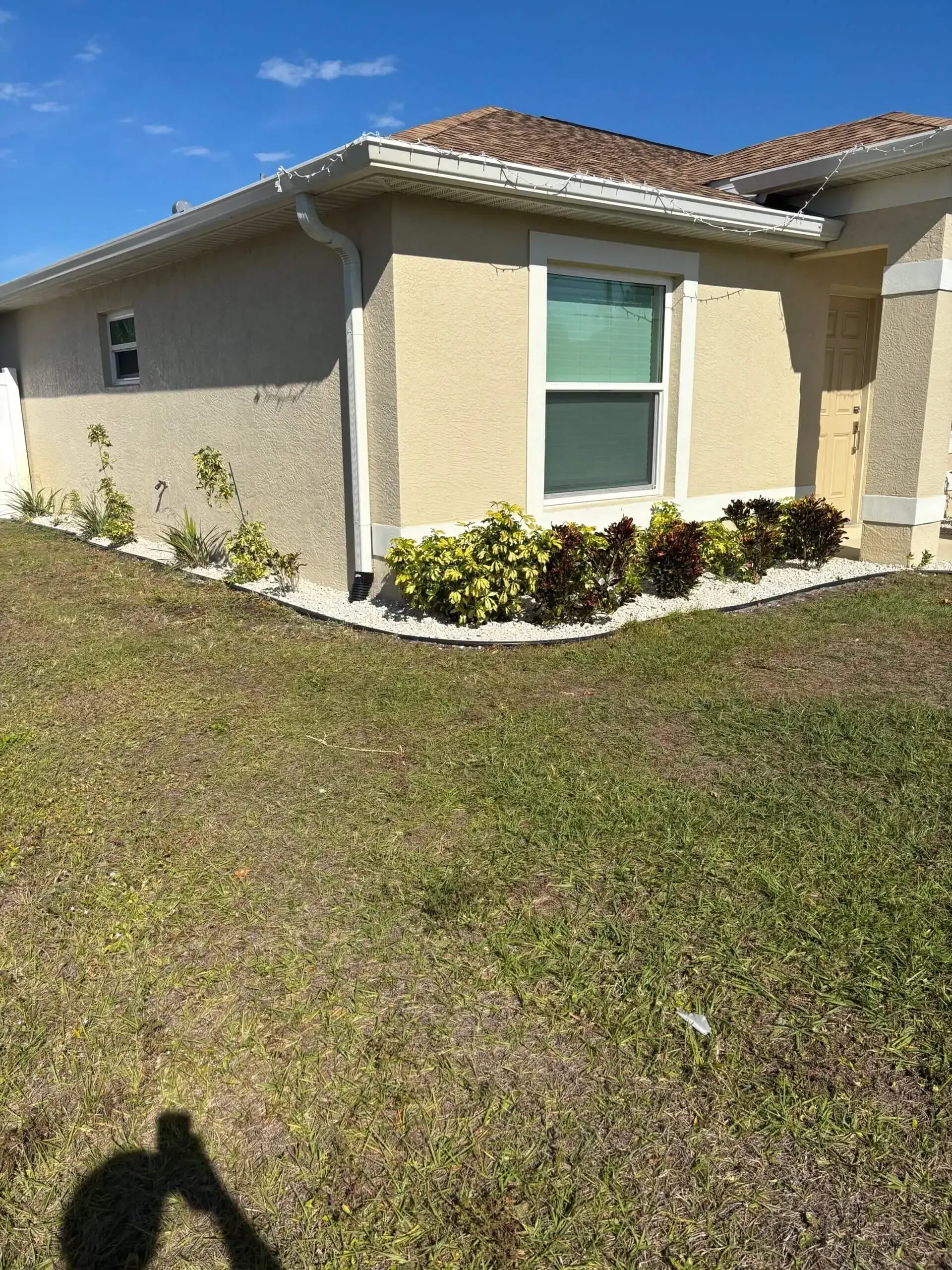 Tan house with white trim and brown roof. Landscaped yard with grass and plants.
