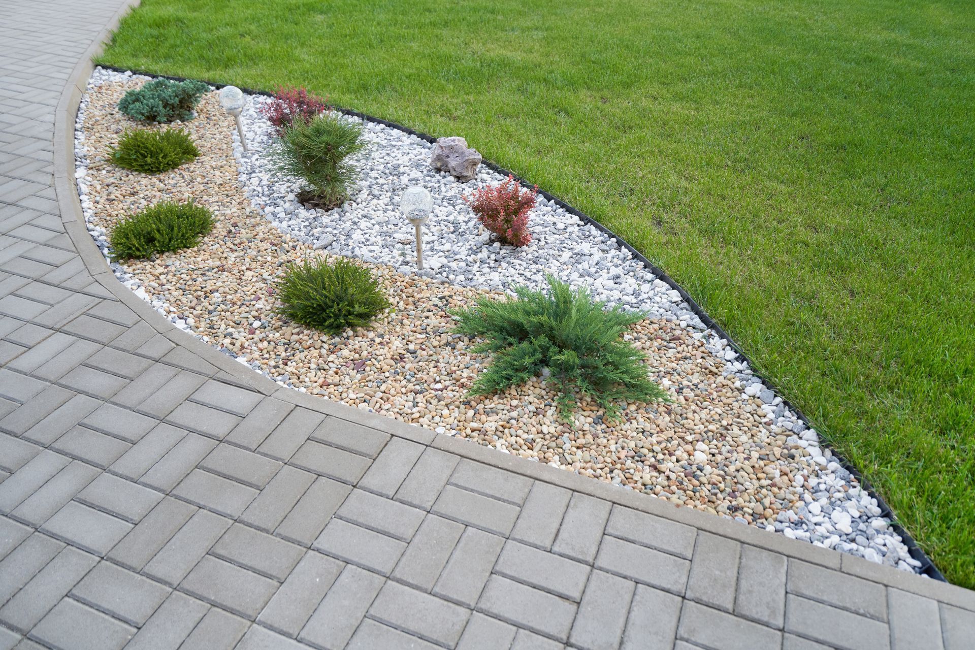 Curved gravel landscape bed with small shrubs beside a gray brick walkway and green lawn