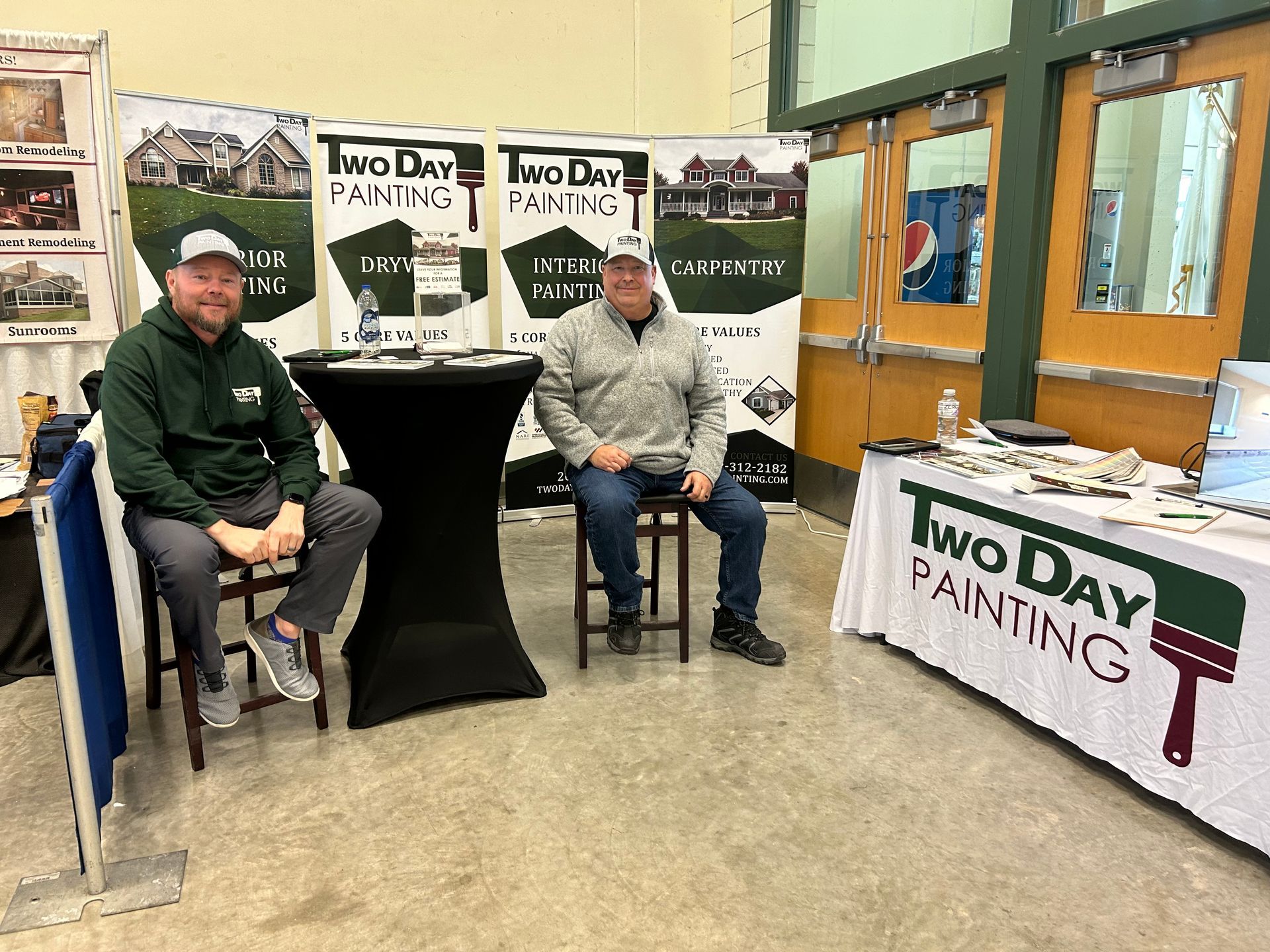 Two men are sitting at a table in front of a two day painting booth.