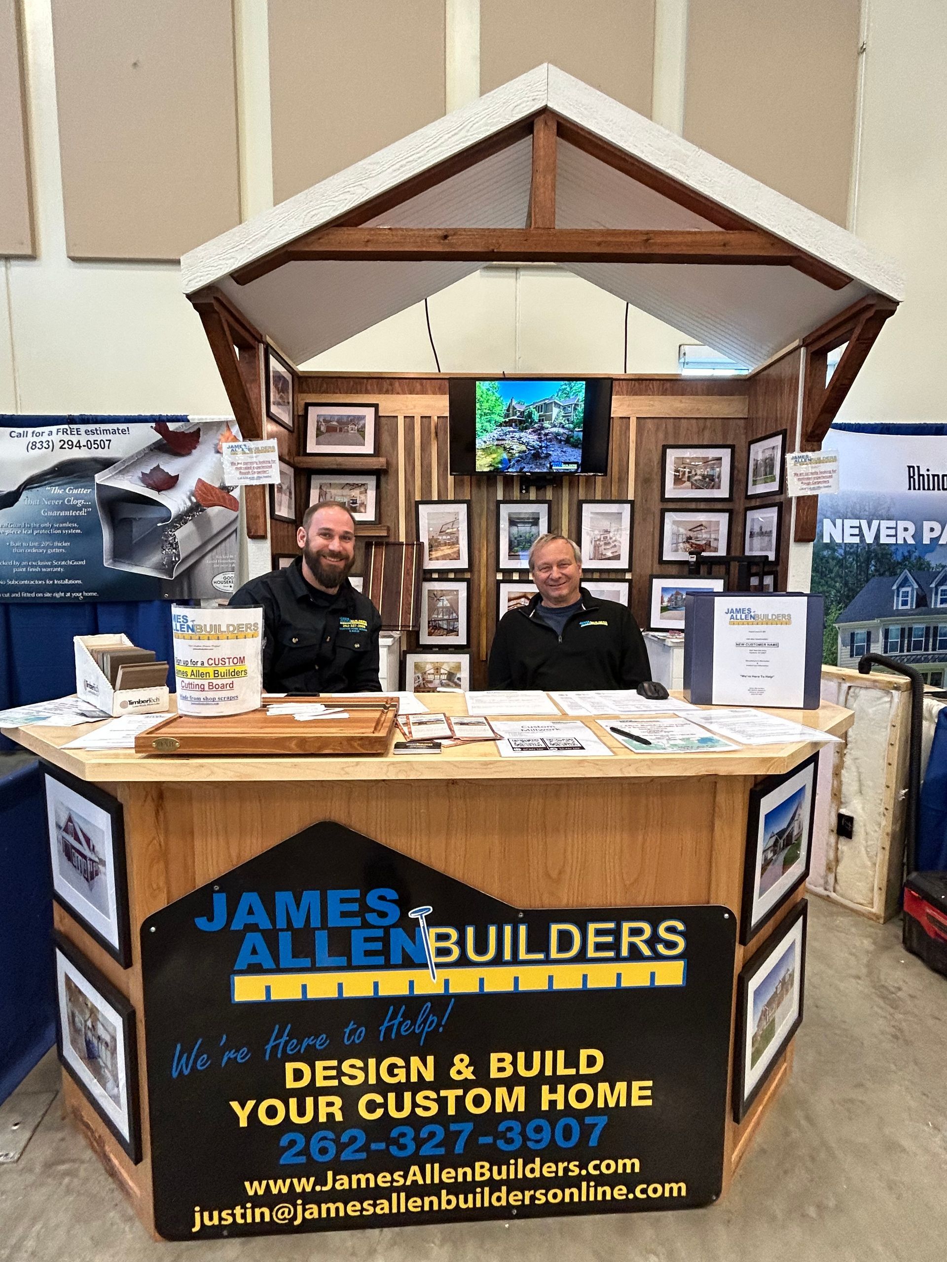 Two men are sitting at a table in front of a james allen builders booth.