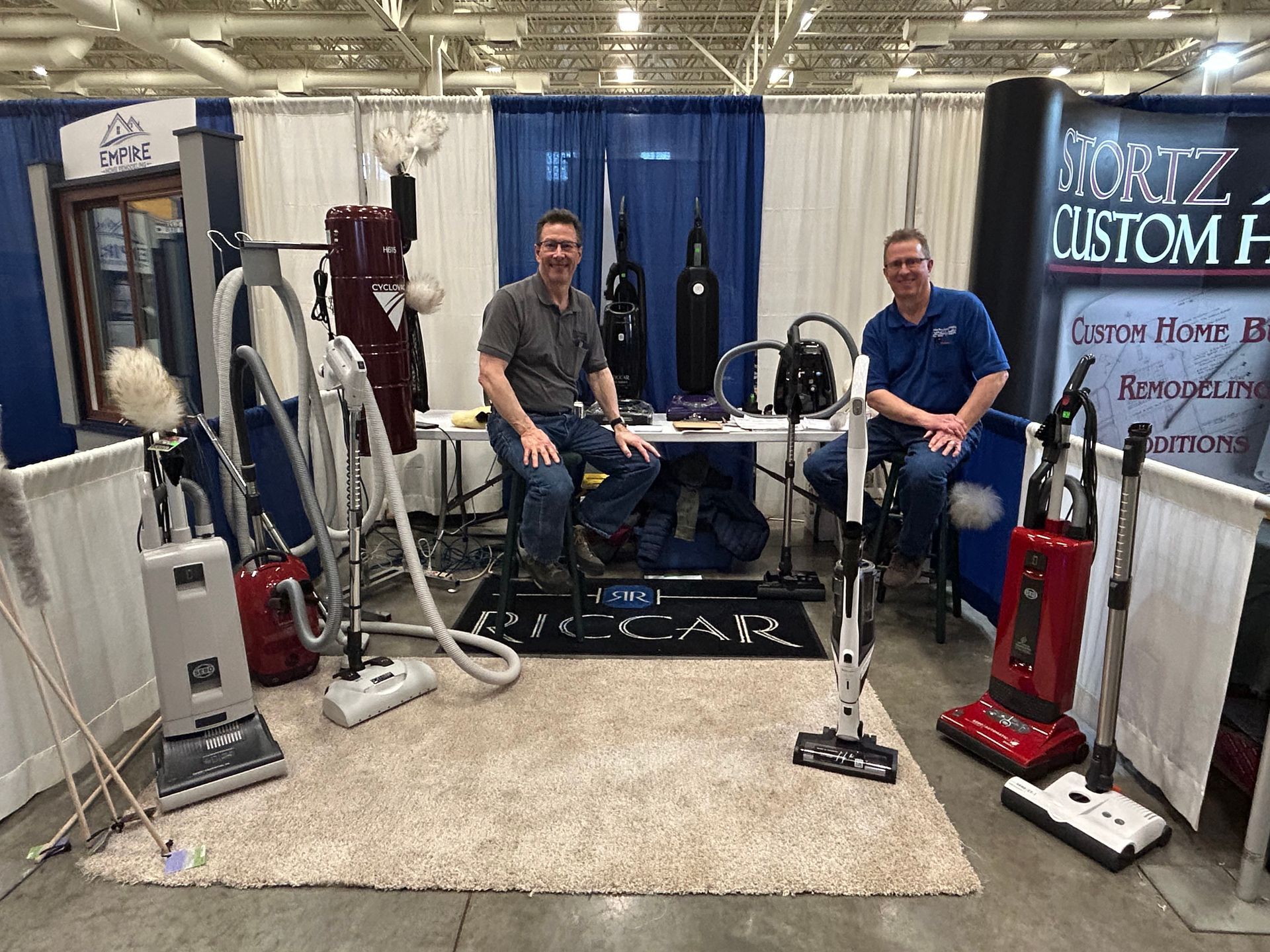 Two men are sitting in front of a vacuum cleaner display.