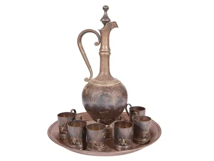Antique silver-colored pitcher with handle and six matching cups on a tray.