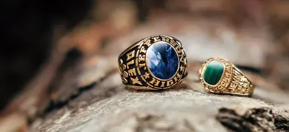 Two gold rings with gemstone settings rest on a textured surface. One has a blue stone, the other green.