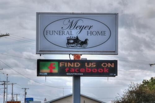 Sign for Meyer Funeral Home, white with black text and carriage illustration, Facebook logo, and text 