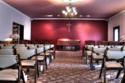 Interior of a chapel with a burgundy wall, wooden chairs, casket, and a cross.
