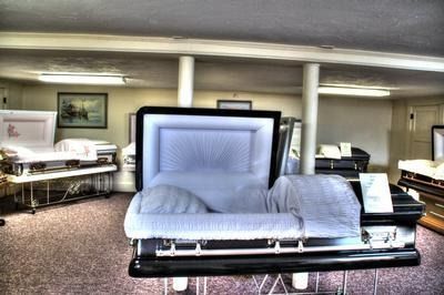 Open black casket in a showroom, surrounded by other caskets. Blue interior, white lighting, and carpet.