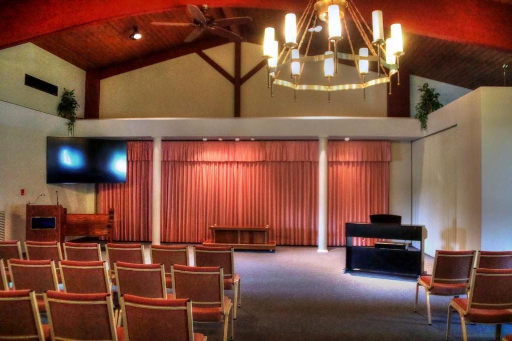 Interior of a chapel with rows of chairs facing a stage with curtains, a large chandelier, and a TV.
