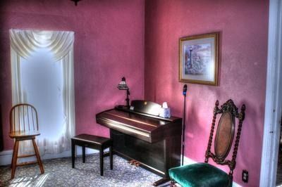Pink-walled room with a piano, two chairs, and a framed picture. Window with white curtains.