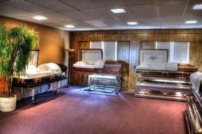 Funeral home interior, multiple open caskets on display, a potted tree, and reddish carpet.