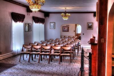 Empty chapel with rows of chairs, chandeliers, and framed art; light streaming through windows.