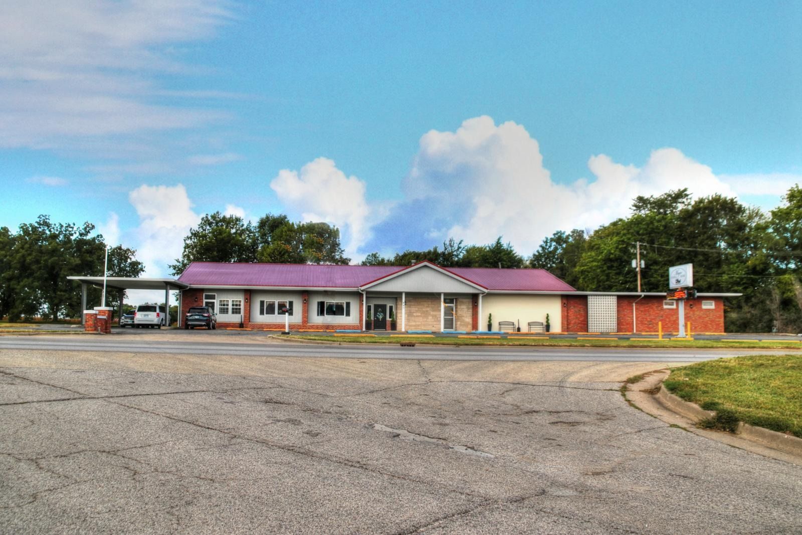 A one-story building with a purple roof sits on a large, cracked asphalt lot, trees in background, and a cloudy sky.