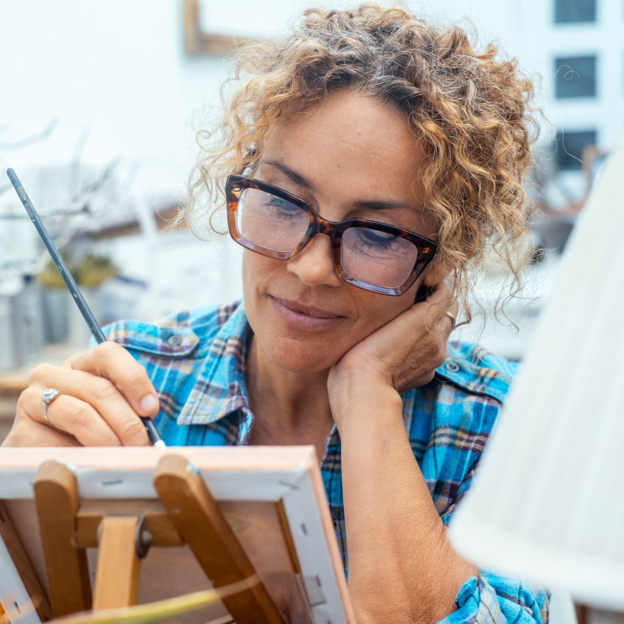 Person sketching on a canvas at an easel, wearing glasses and a blue plaid shirt
