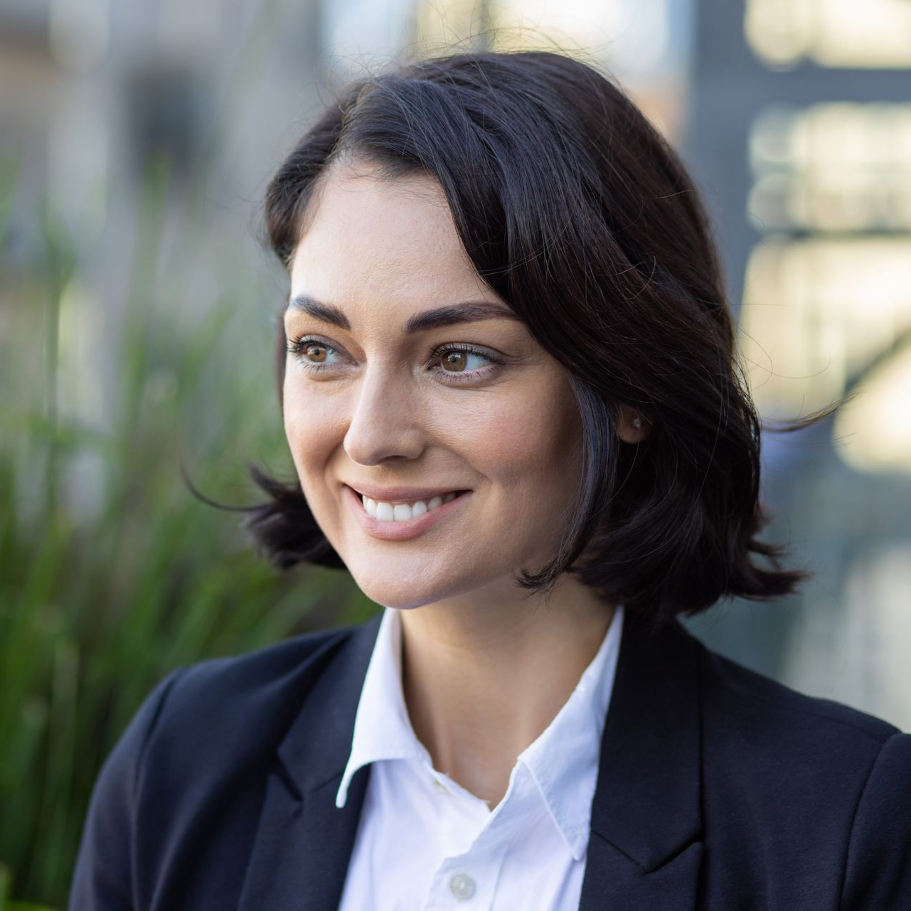 Smiling woman in a dark blazer and white shirt outdoors, with blurred greenery behind her