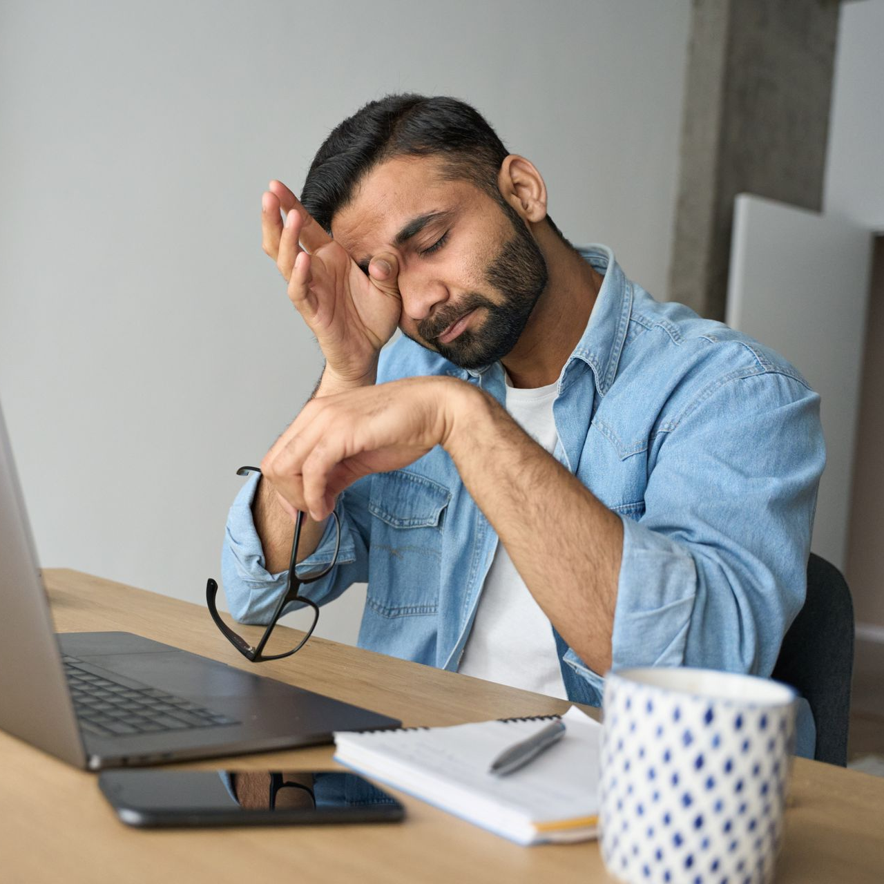 Tired man resting his head on his hand at a desk with a laptop, notebook, and coffee mug
