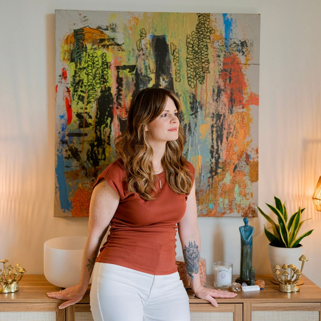 Woman standing by a wooden console in a bright room, with an abstract painting and lamps behind her.