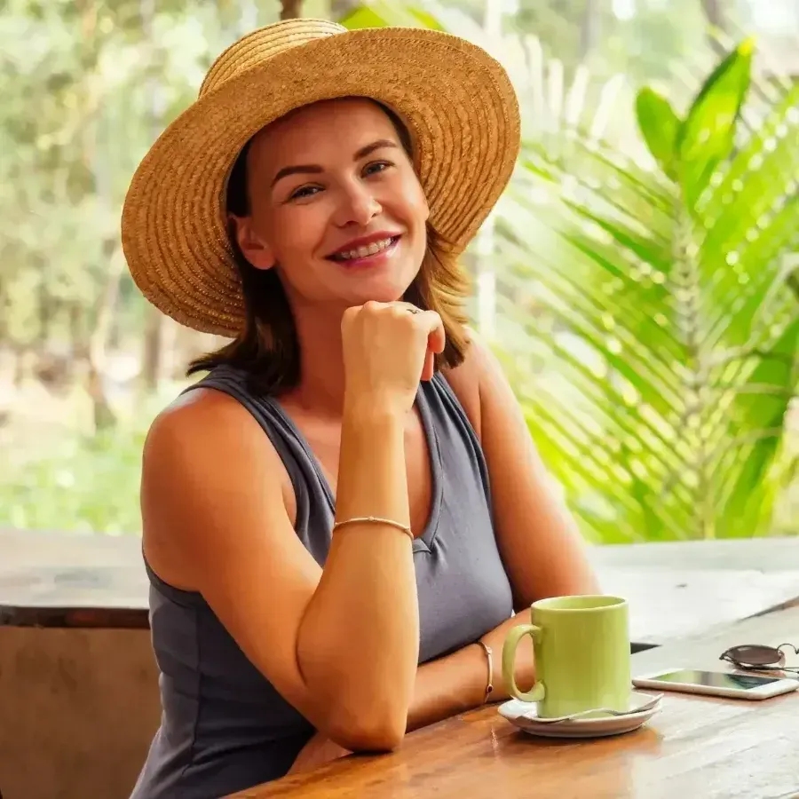 Smiling woman in a straw hat sitting at a table with a green coffee cup on a sunny patio