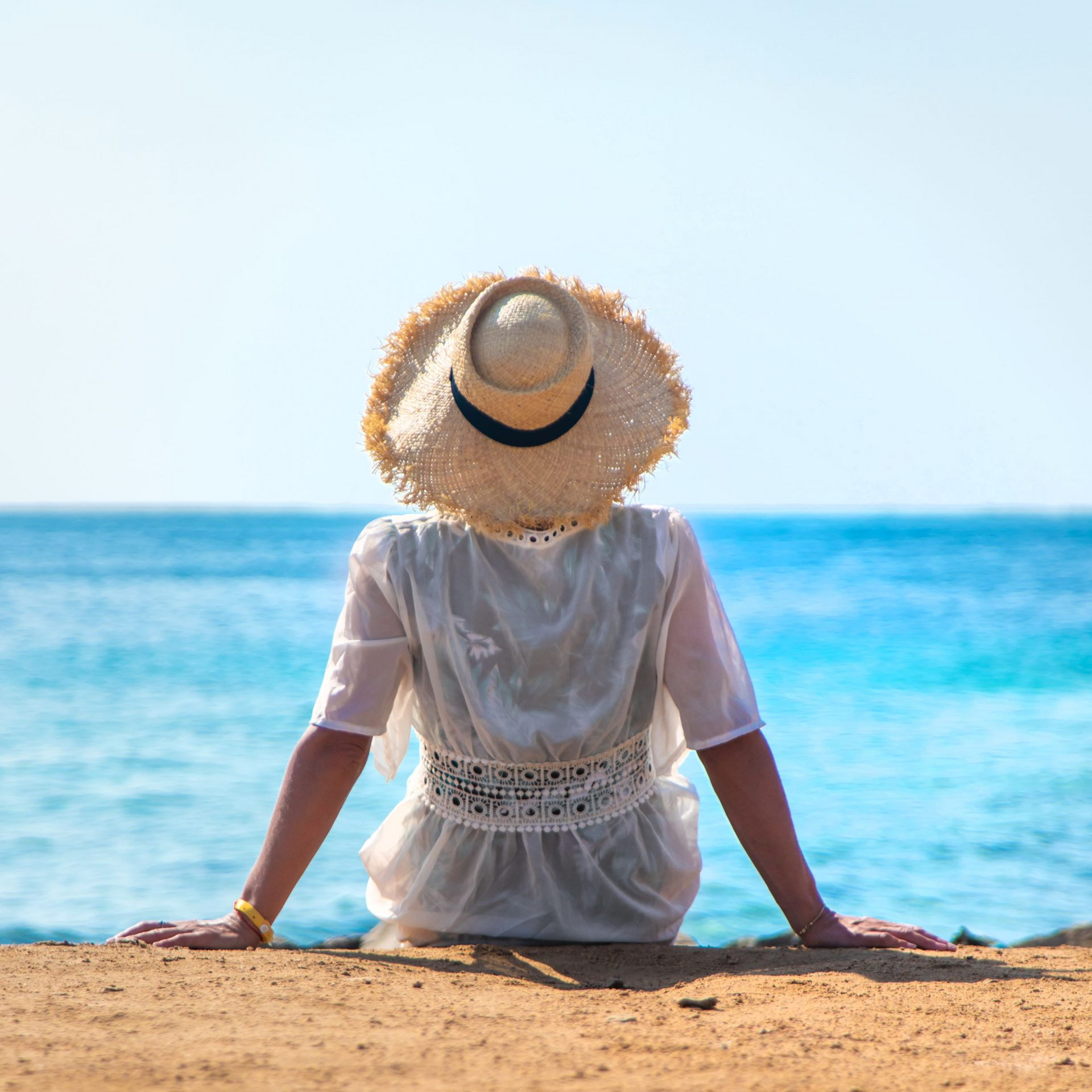 Person sitting on a sandy beach facing the blue ocean, wearing a straw hat and white dress