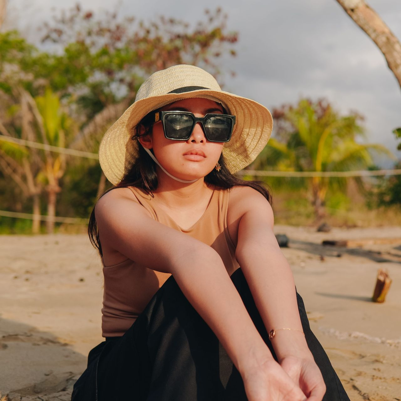 Woman sitting on a sandy beach wearing a straw hat and sunglasses, with palm trees in the background
