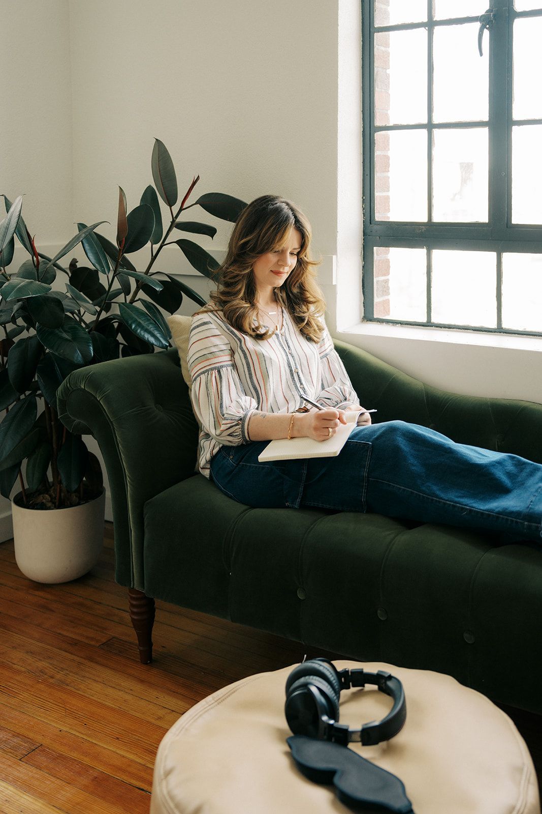 Woman reclining on a green chaise by a window, reading a book with headphones on a nearby table.