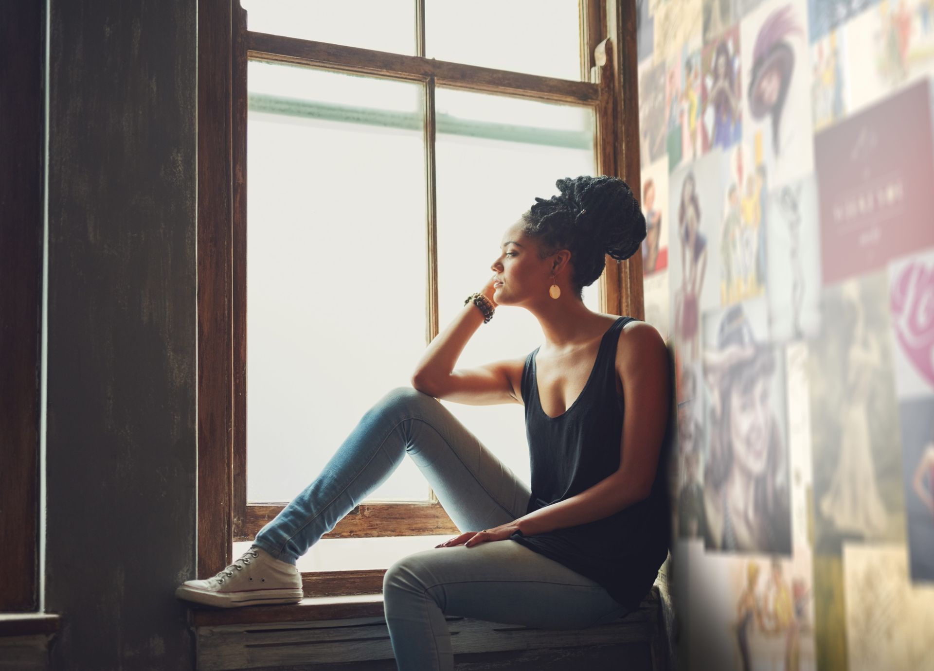 Person sitting on a window ledge, looking out thoughtfully in a dim room with posters on the wall.