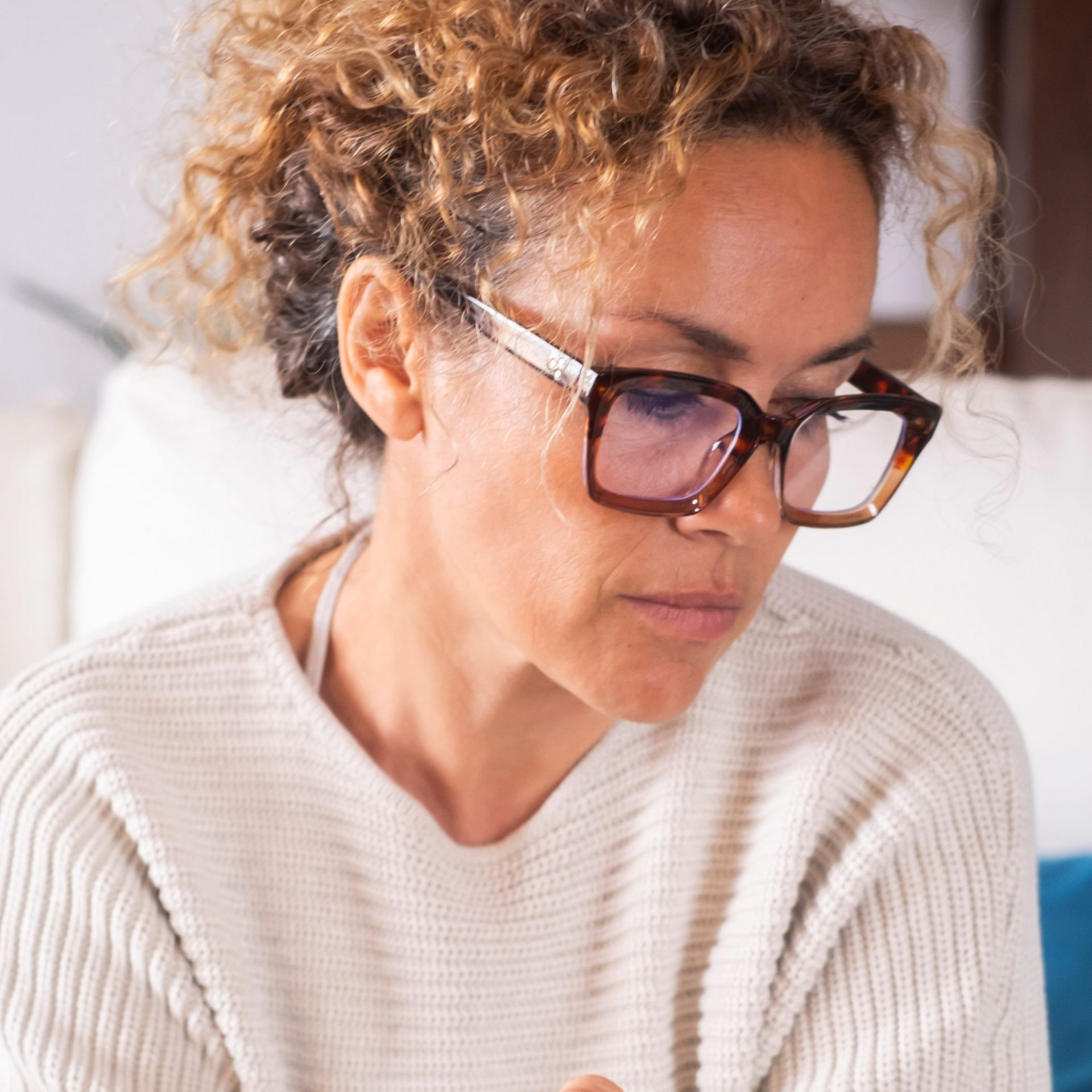 Woman with curly hair and glasses looking down, wearing a cream sweater indoors