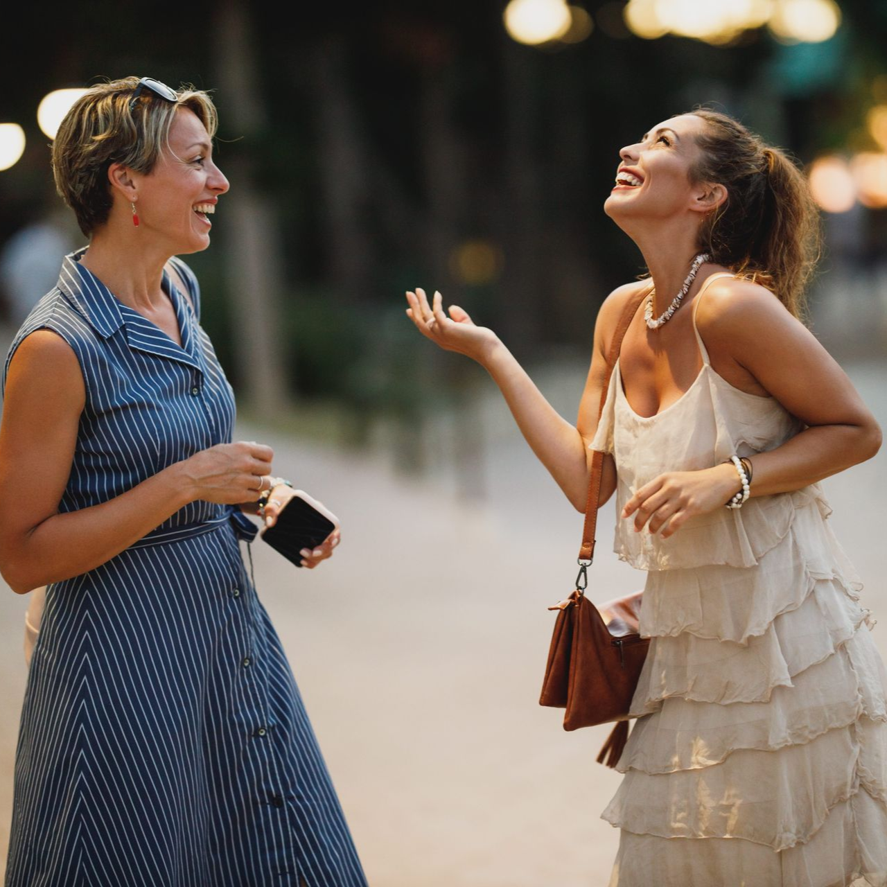 Two women chatting and laughing outdoors, one in a blue dress and one in a cream ruffled dress.