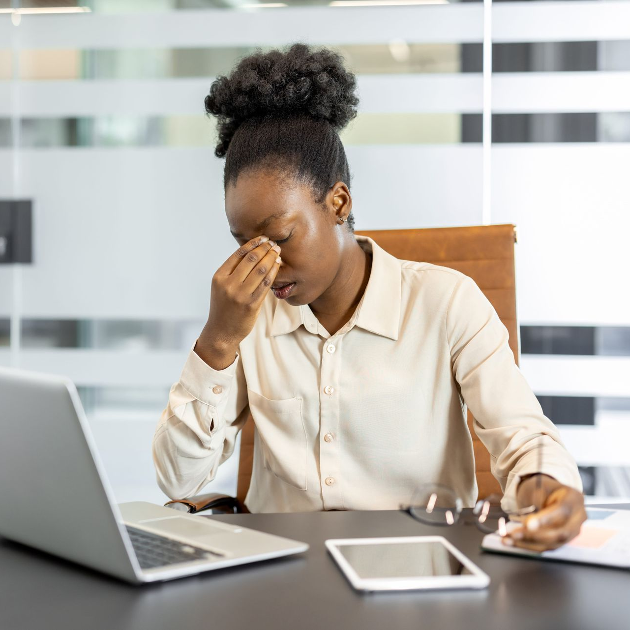 Woman at desk rubbing her eyes beside a laptop and tablet, looking tired in a bright office