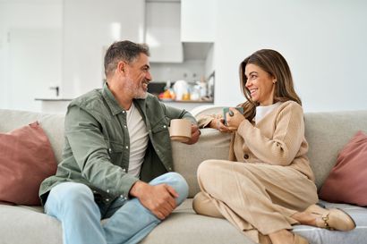 Two people sitting on a couch, smiling and toasting mugs in a bright living room.