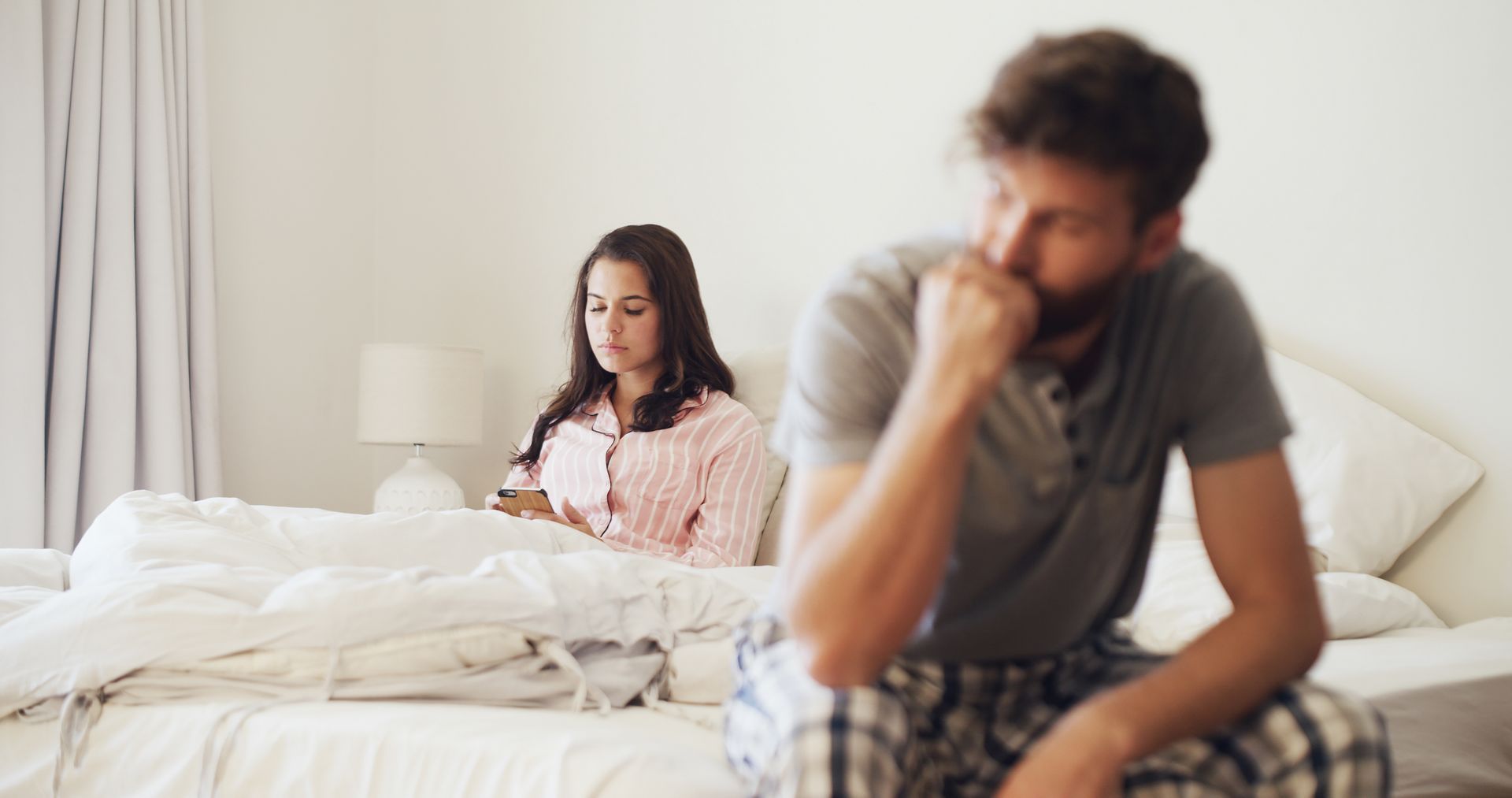 A couple sits apart on a bed, each looking upset in a bright bedroom.