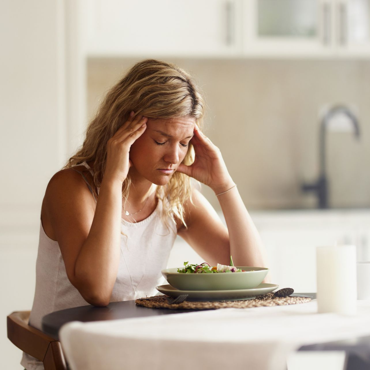 Person sitting at a table with head in hands beside a plate of salad in a bright kitchen