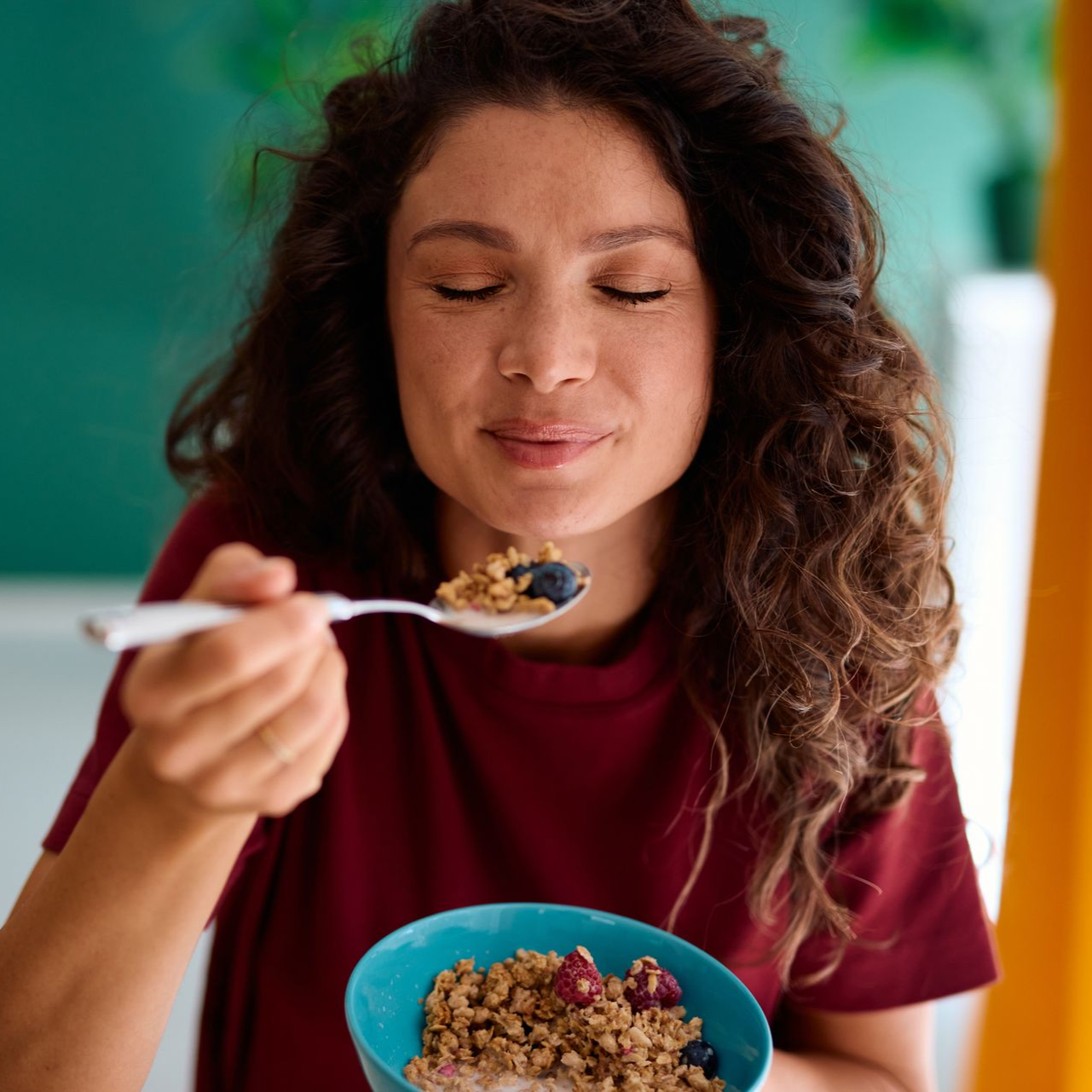Woman eating cereal from a spoon beside a blue bowl, eyes closed, in a bright kitchen