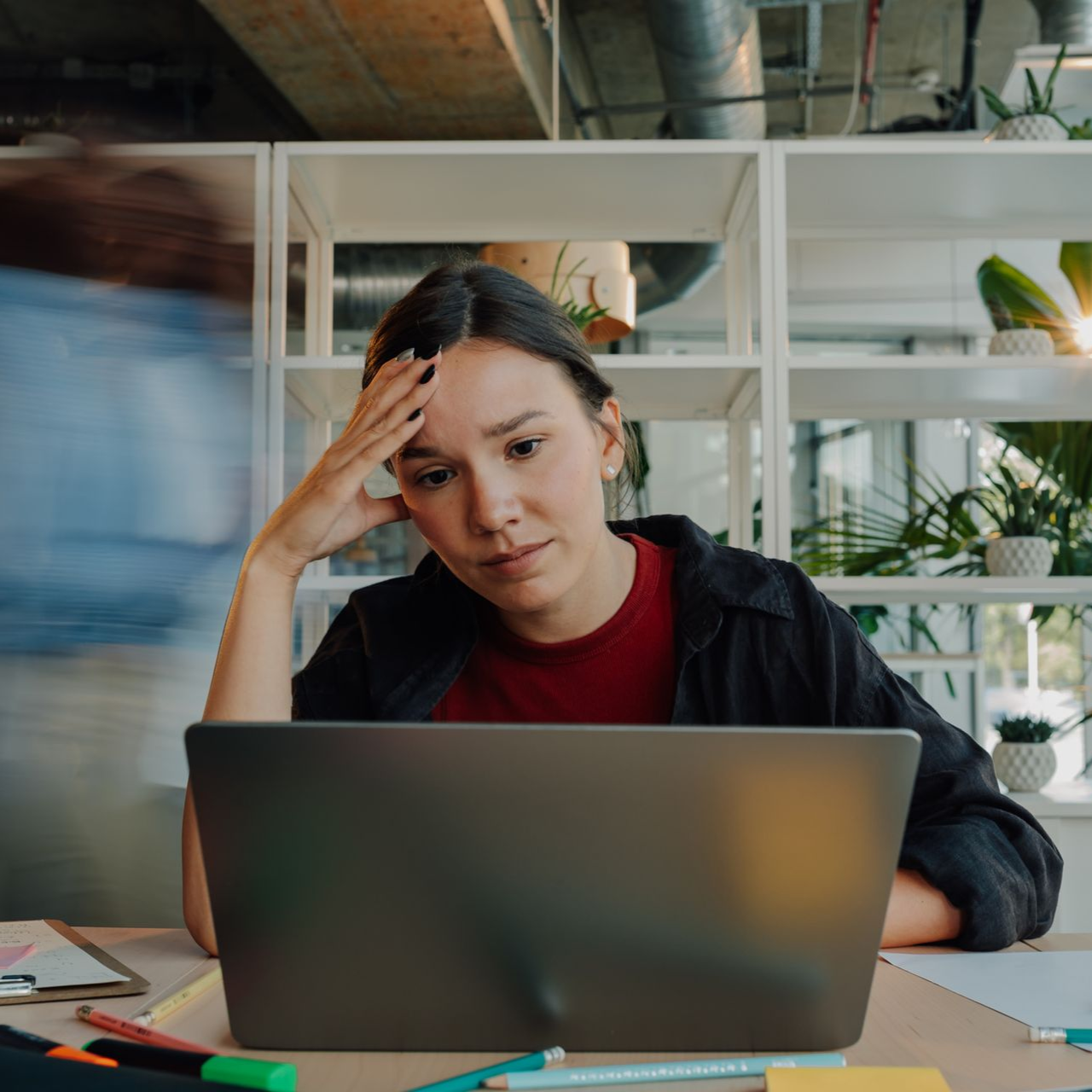 Person stressed at a laptop in a bright office, hand on forehead while working