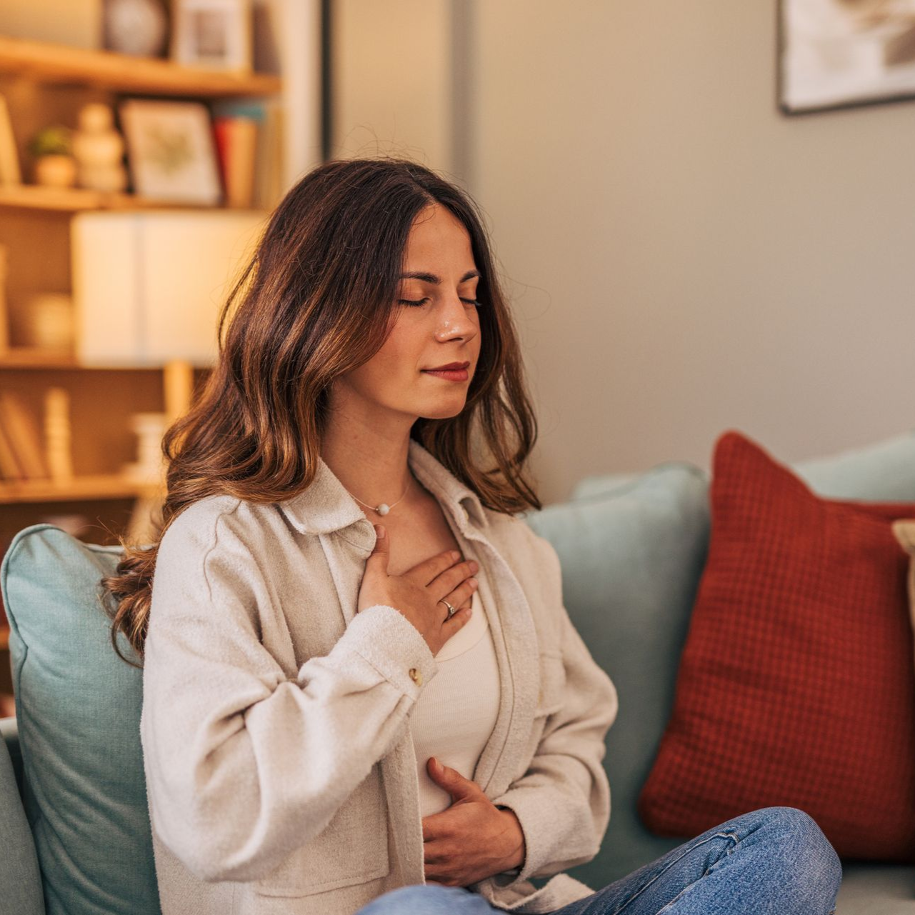 Woman meditating on a sofa in a cozy living room, with hand on chest and eyes closed