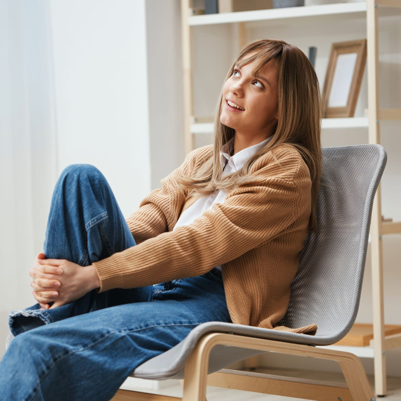 Woman sitting in a chair by a bookshelf, wearing a tan sweater and jeans, looking upward thoughtfully