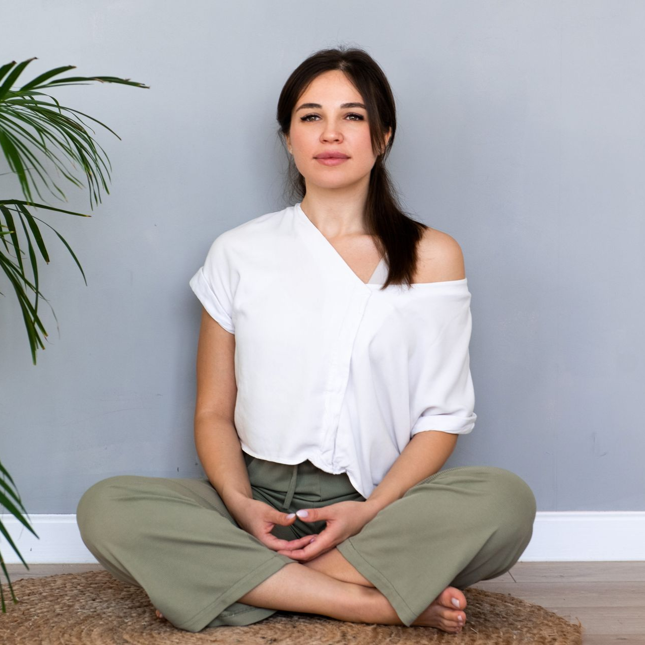 Woman sitting cross-legged on the floor, meditating in a bright room with a potted plant.