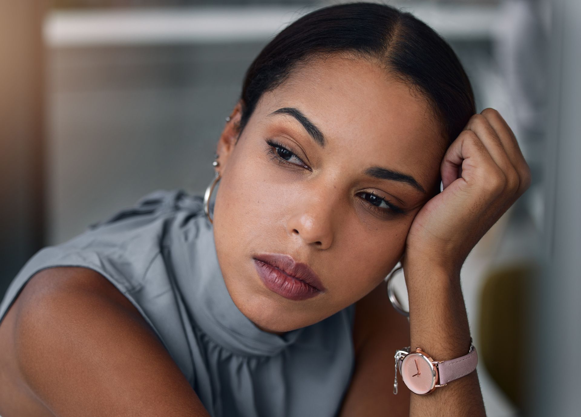Woman in gray top resting her head on her hand, looking thoughtful indoors.