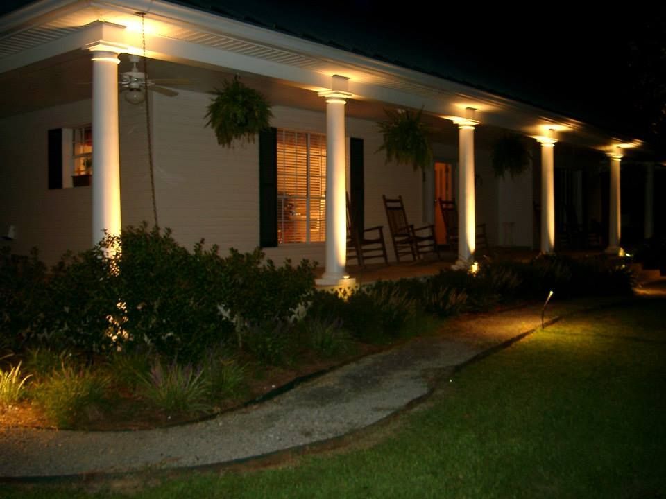 A house porch at night with pillars illuminated by lights, hanging plants, and a path leading to the entrance.