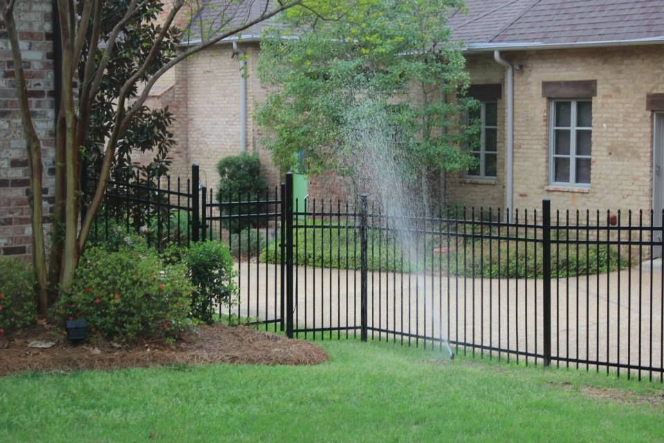 A spray of water from a garden sprinkler shoots over a black metal fence in front of a brick house.