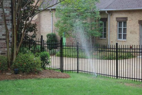 A spray of water from a garden sprinkler shoots over a black metal fence in front of a brick house.