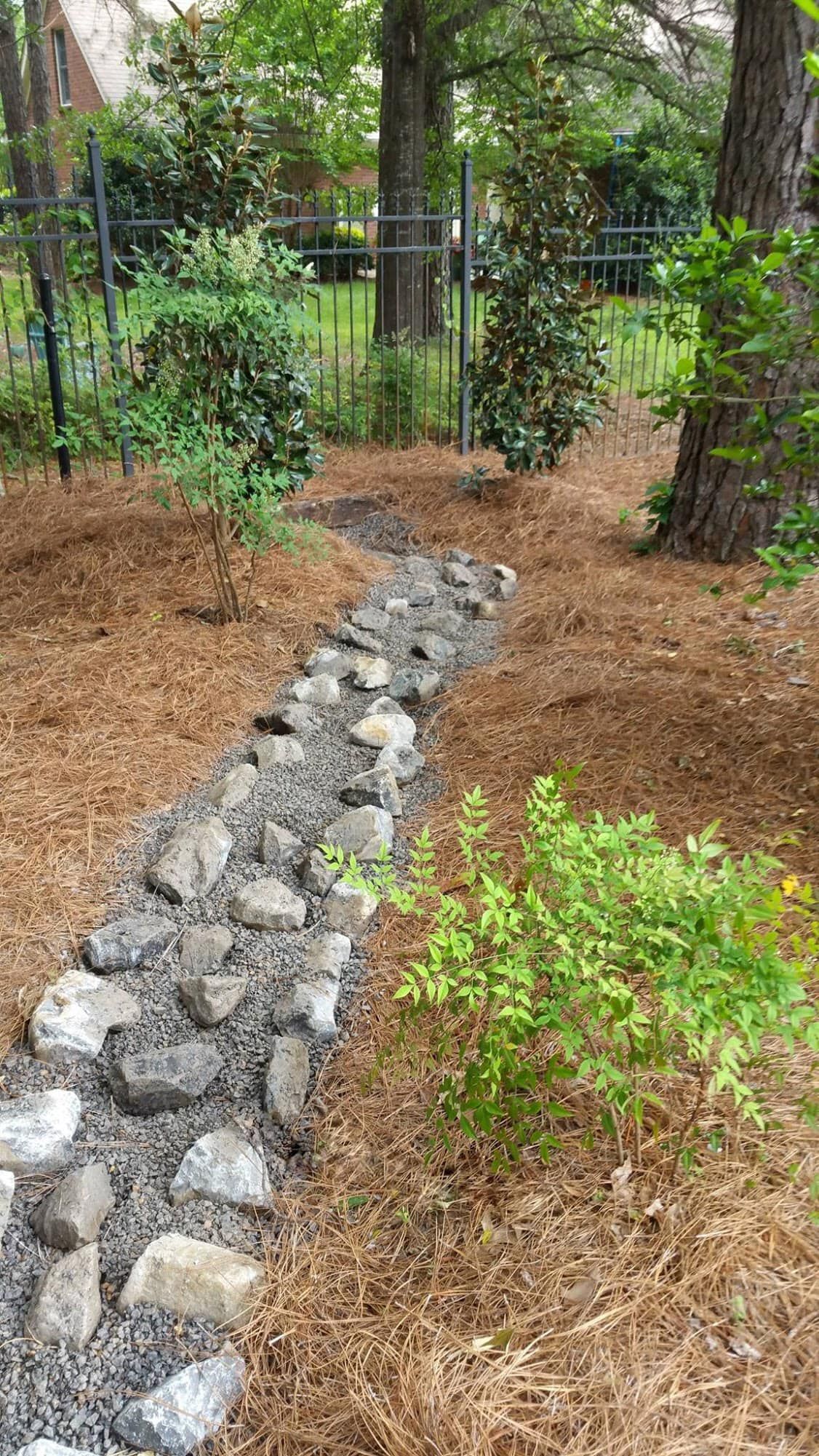 A dry creek bed made of gray river rocks winds through a mulched garden area with surrounding shrubs and trees.