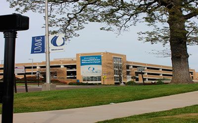 A tan parking garage with blue UMMC banners and signs under a clear sky next to a large tree and a concrete walkway.