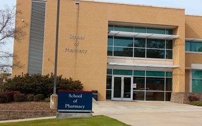 A tan brick School of Pharmacy building with a blue sign out front and glass entry doors on a sunny day.