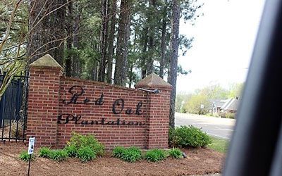 A brick sign with Red Oak Plantation in dark script lettering, situated in a wooded area next to a paved road.