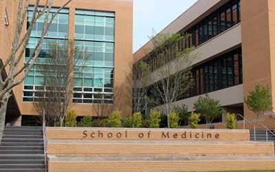 The tan brick building features a School of Medicine sign on its front steps, flanked by stairs and small trees.