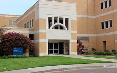 The tan brick building of a School of Nursing, featuring a central white entrance with a decorative arched window.
