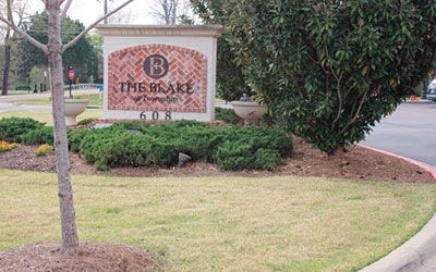 A stone monument sign for The Blake at 608, featuring brickwork, landscaping, and a tree in front of a paved driveway.