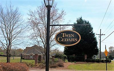 An oval sign reading Twin Cedars hangs from a black metal post in front of a residential area with trees and a lawn.